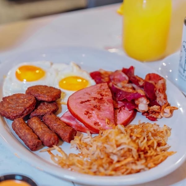 A full breakfast plate with eggs, sausage, bacon, ham, hash browns, and orange juice.