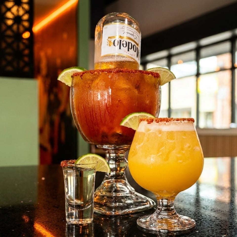 A large Michelada with a Modelo bottle, a shot of tequila, and a yellow drink on a bar.
