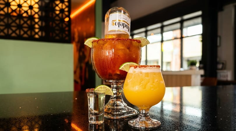 A large michelada with a Modelo beer, a yellow drink, and a tequila shot on a bar.