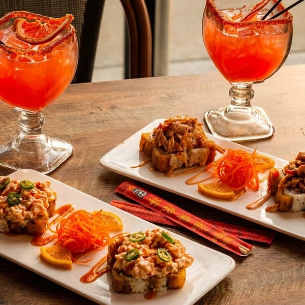 Two plates of sushi rolls with orange slices and two red cocktails on a wooden table.