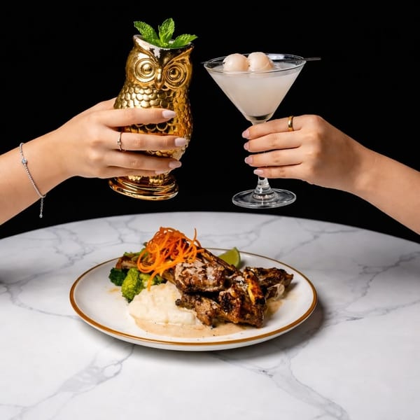 Two hands clinking a golden owl cocktail and a lychee martini over a plate of lamb chops.